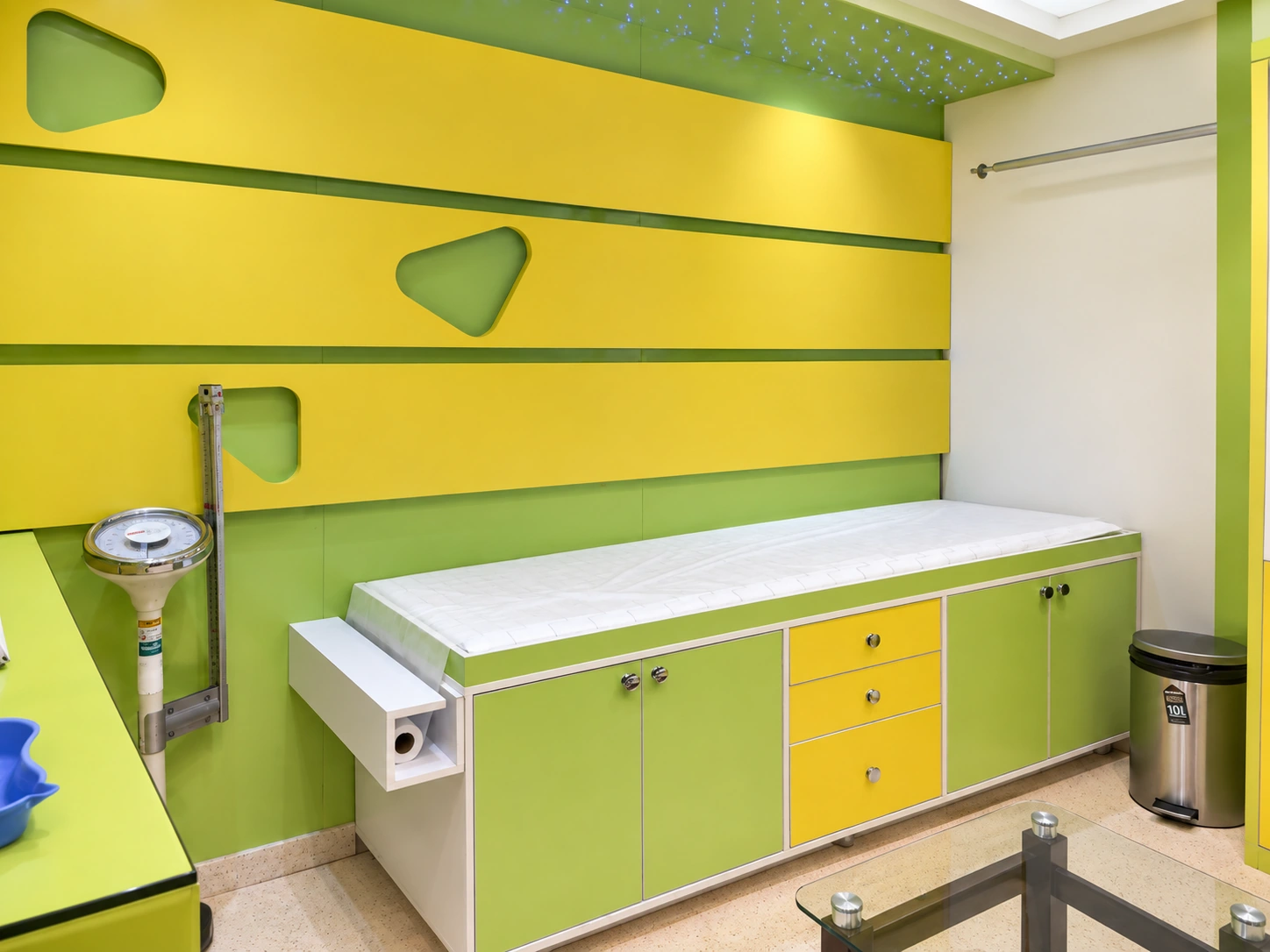 Pediatric examination table area at Day Hospital with green and yellow wall panels, built-in storage and clinical measuring equipment