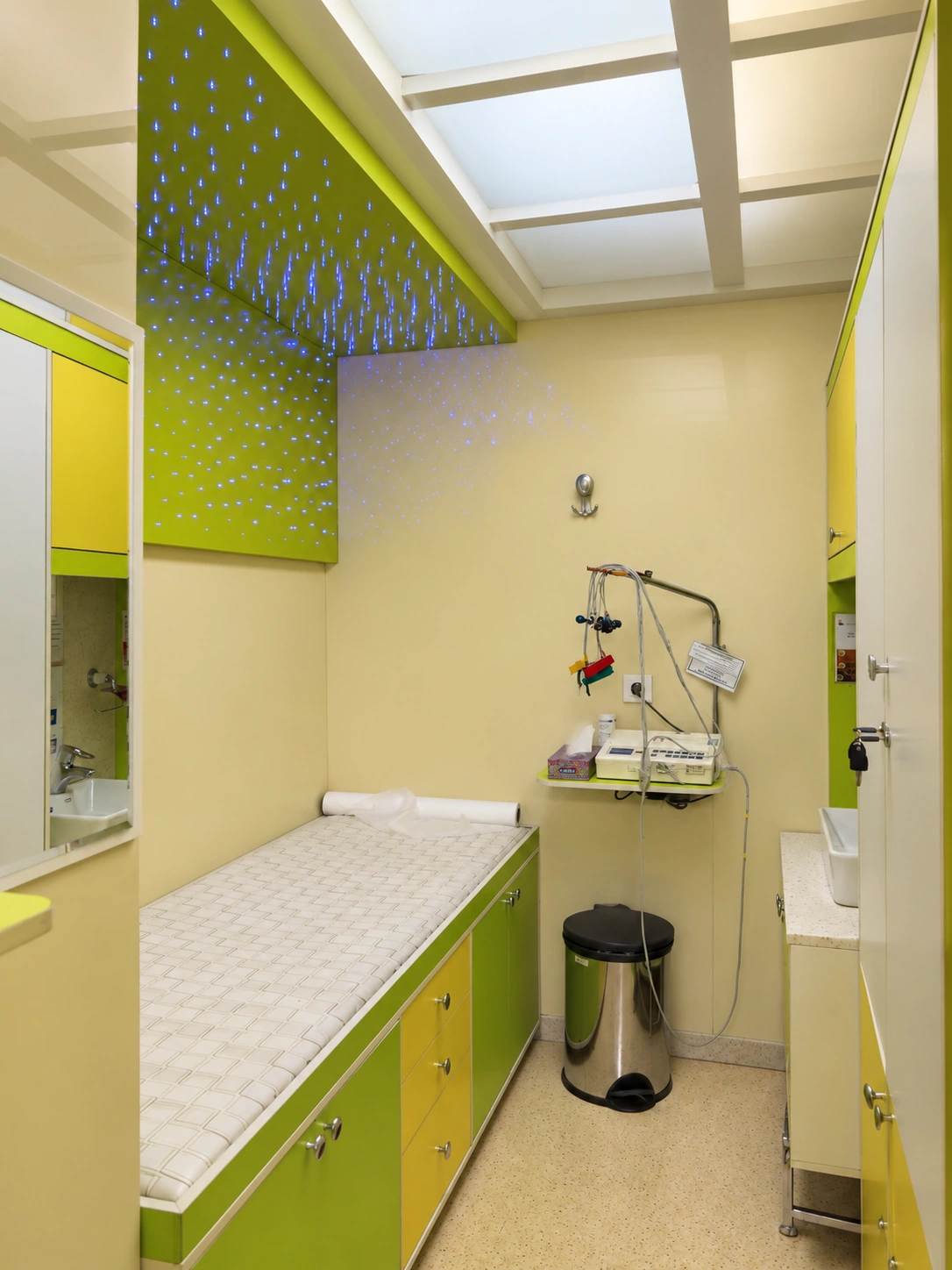 Day Hospital pediatric treatment room with examination table, integrated green-yellow cabinetry, luminous ceiling panels and star-like blue lighting